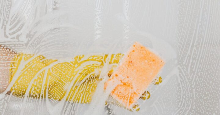 Hand with yellow glove holding sponge cleaning glass surface.