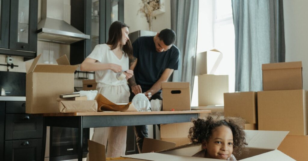 A mom and dad preparing for a move in their kitchen while their daughter is playing in a cardboard moving box.