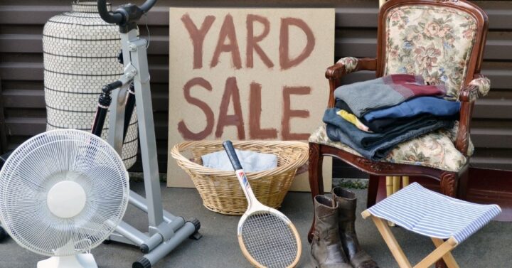 A Yard Sale sign surrounded by a small white fan, tennis racket, floral arm chair, folded up pile of clothing, and a lamp