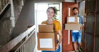 A college-aged man and woman carrying boxes labeled, cups and books, into an apartment complex.