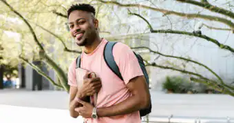 A college-aged man holding a book while wearing a book bag on what appears to be a college campus.