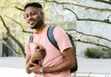 A college-aged man holding a book while wearing a book bag on what appears to be a college campus.