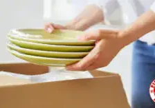 A person carefully unpacking ceramic dishes from a moving box in their new kitchen, featured in a guide on how to find affordable movers.