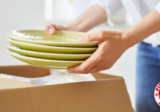 A person carefully unpacking ceramic dishes from a moving box in their new kitchen, featured in a guide on how to find affordable movers.